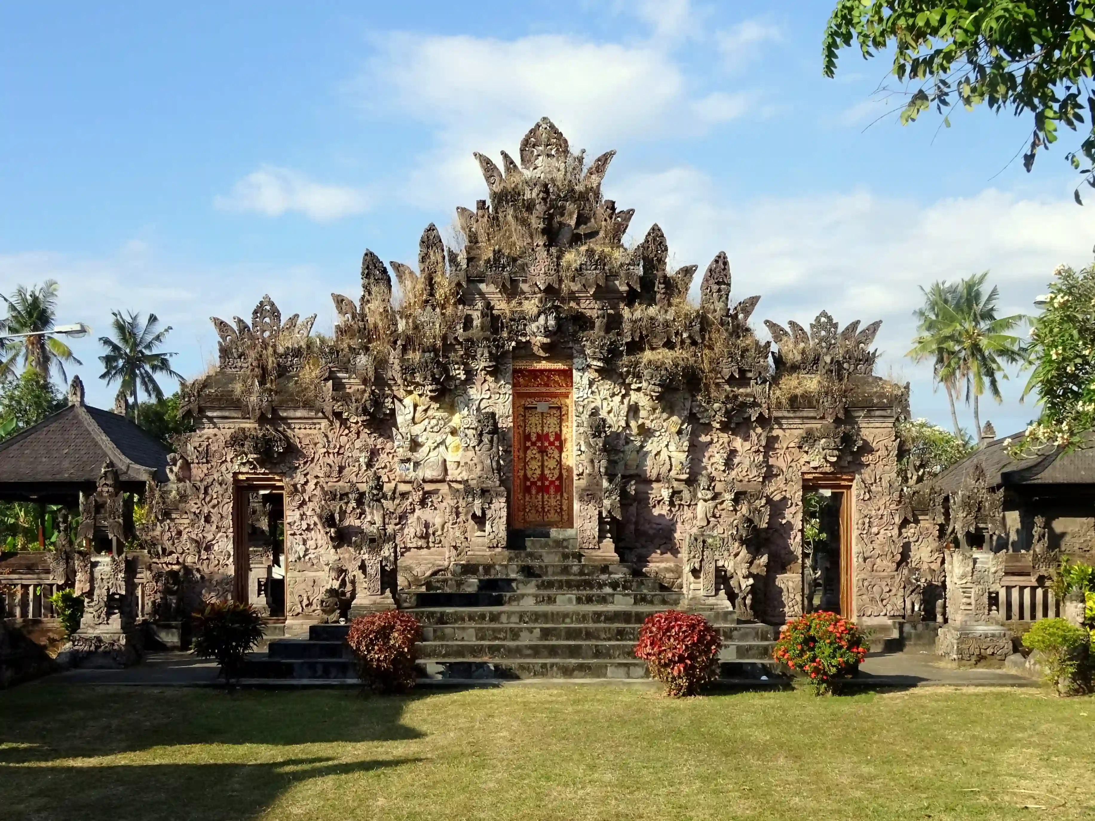 Front view of a highly decorated, carved stone Balinese temple gate with steps and a red door.