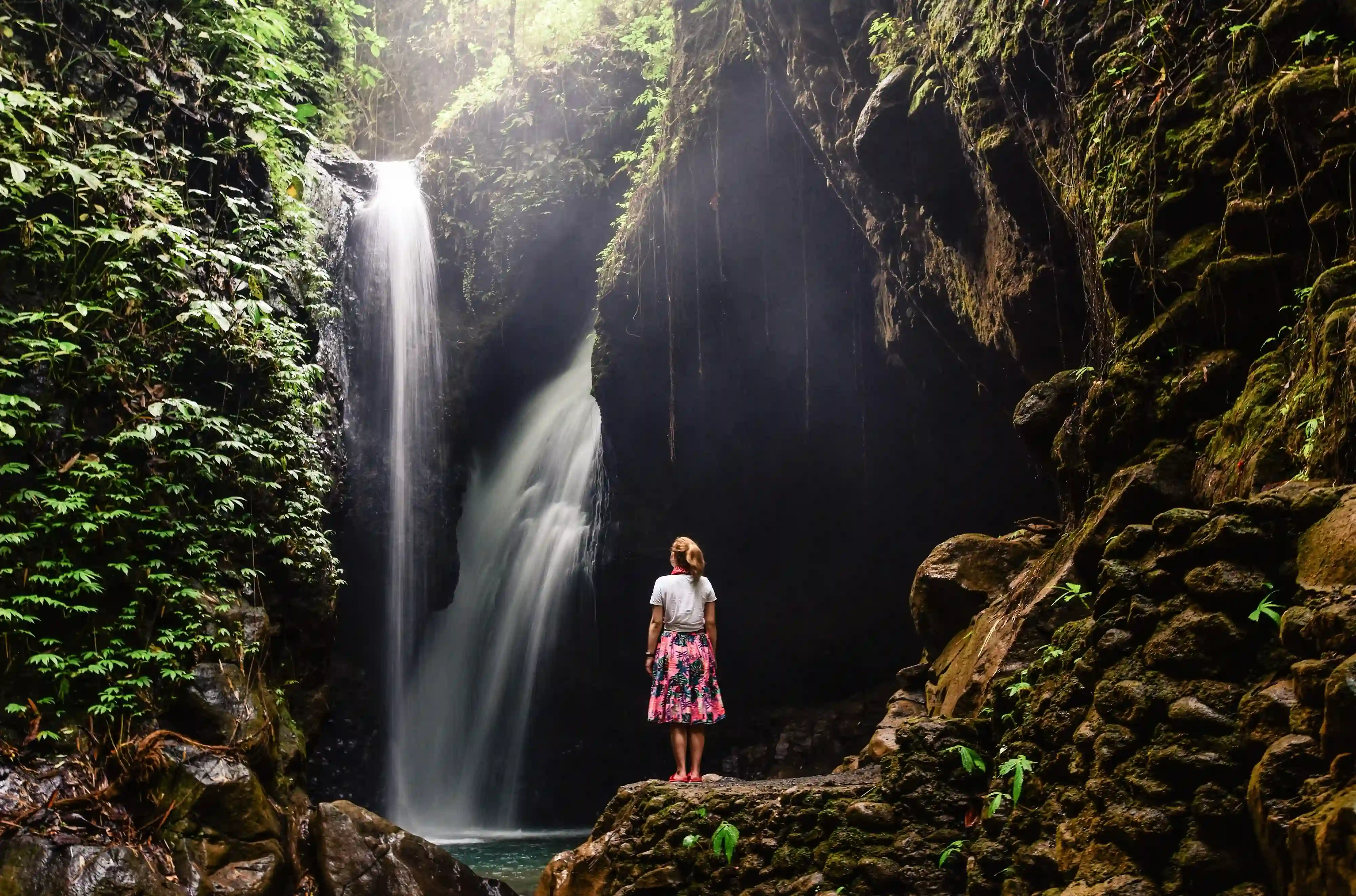Woman standing on a rock looking at a powerful waterfall cascading down into a dark, rocky pool in a lush jungle.