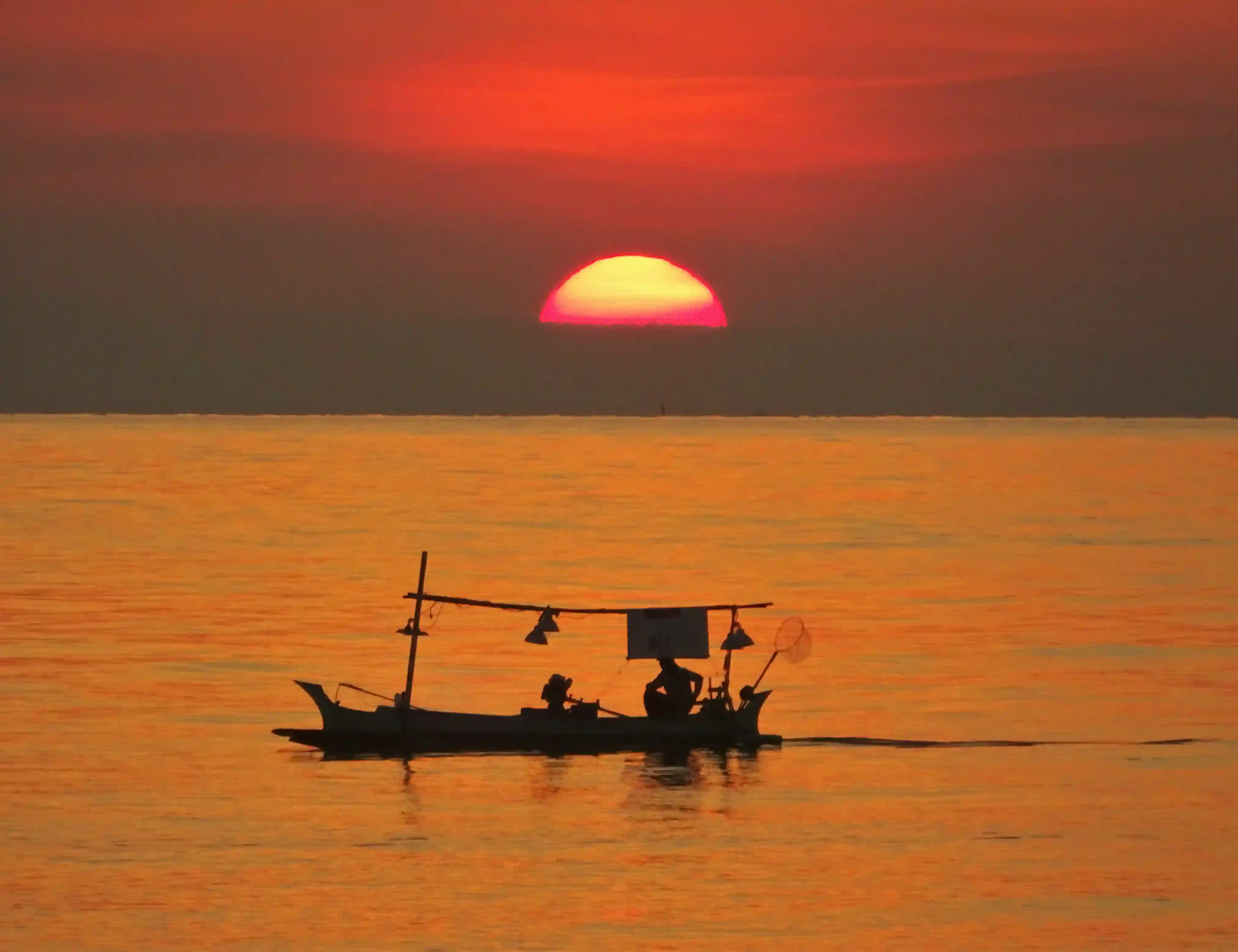 A traditional Balinese Jukung fishing boat silhouetted against a bright orange and red sunset over the ocean.