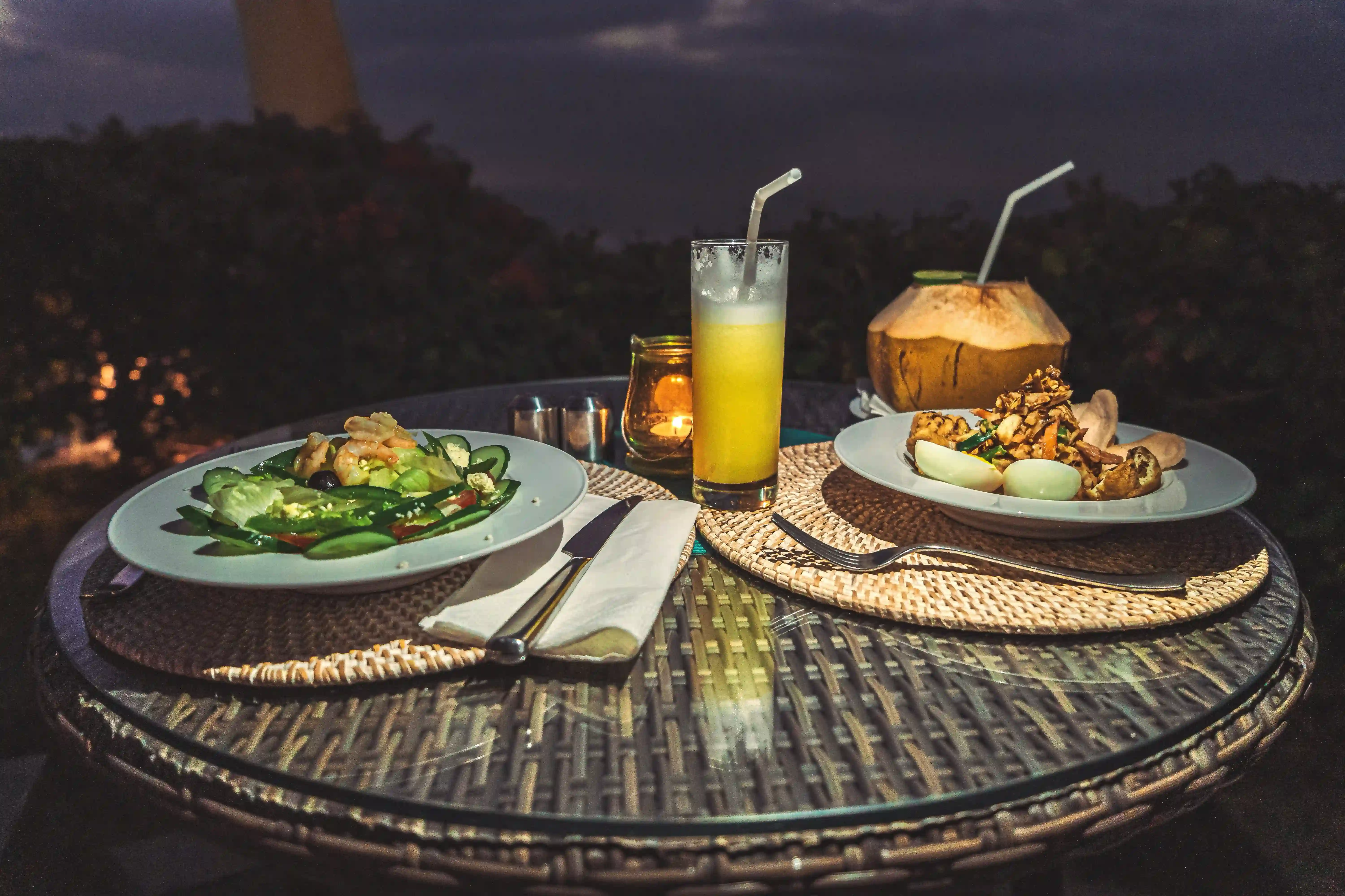 A romantic dinner setting at night with two plates of food, juice, a fresh coconut, and candlelight.