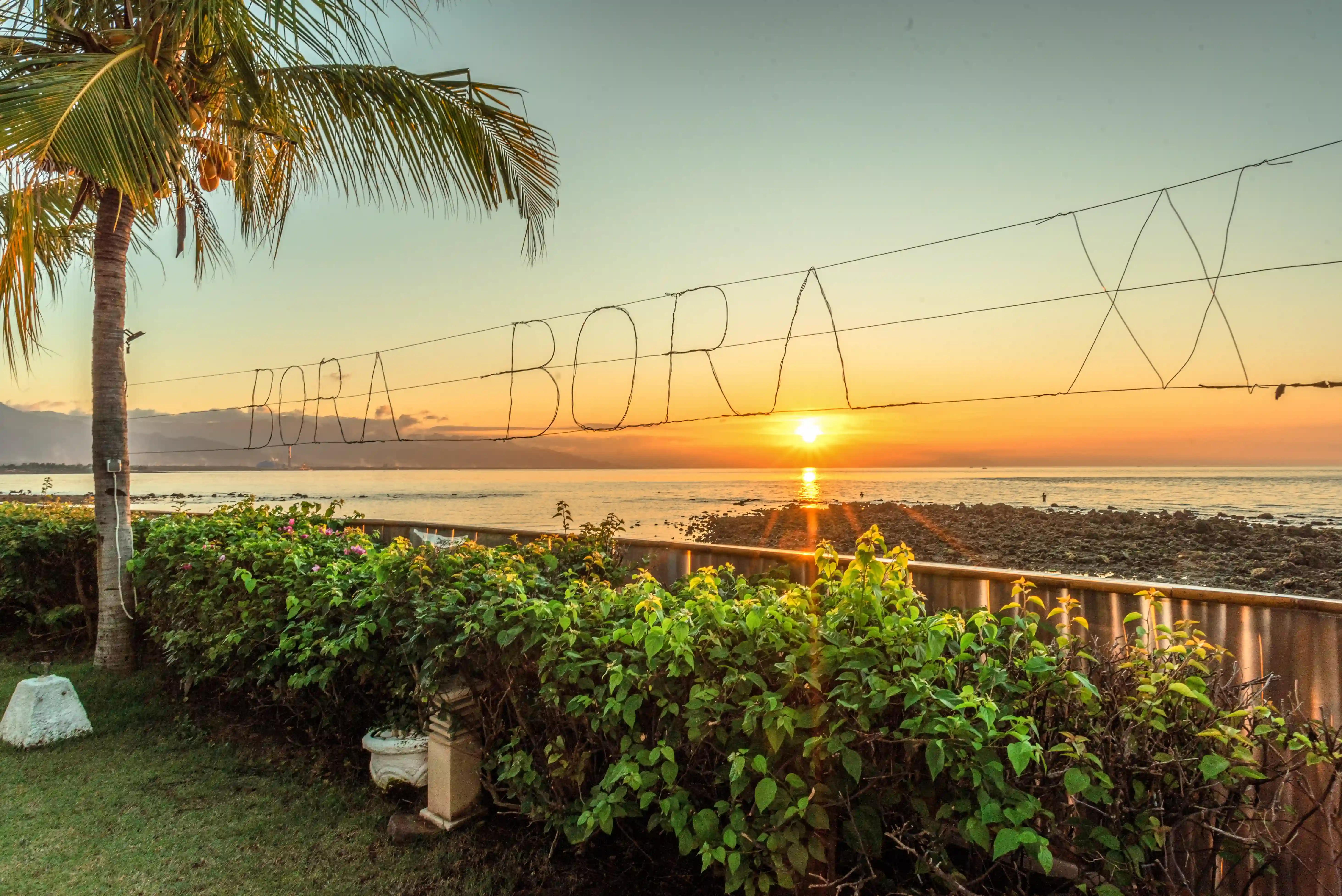 A tropical sunset over the ocean with the restaurant name 'Bora Bora' displayed in lights over the water.