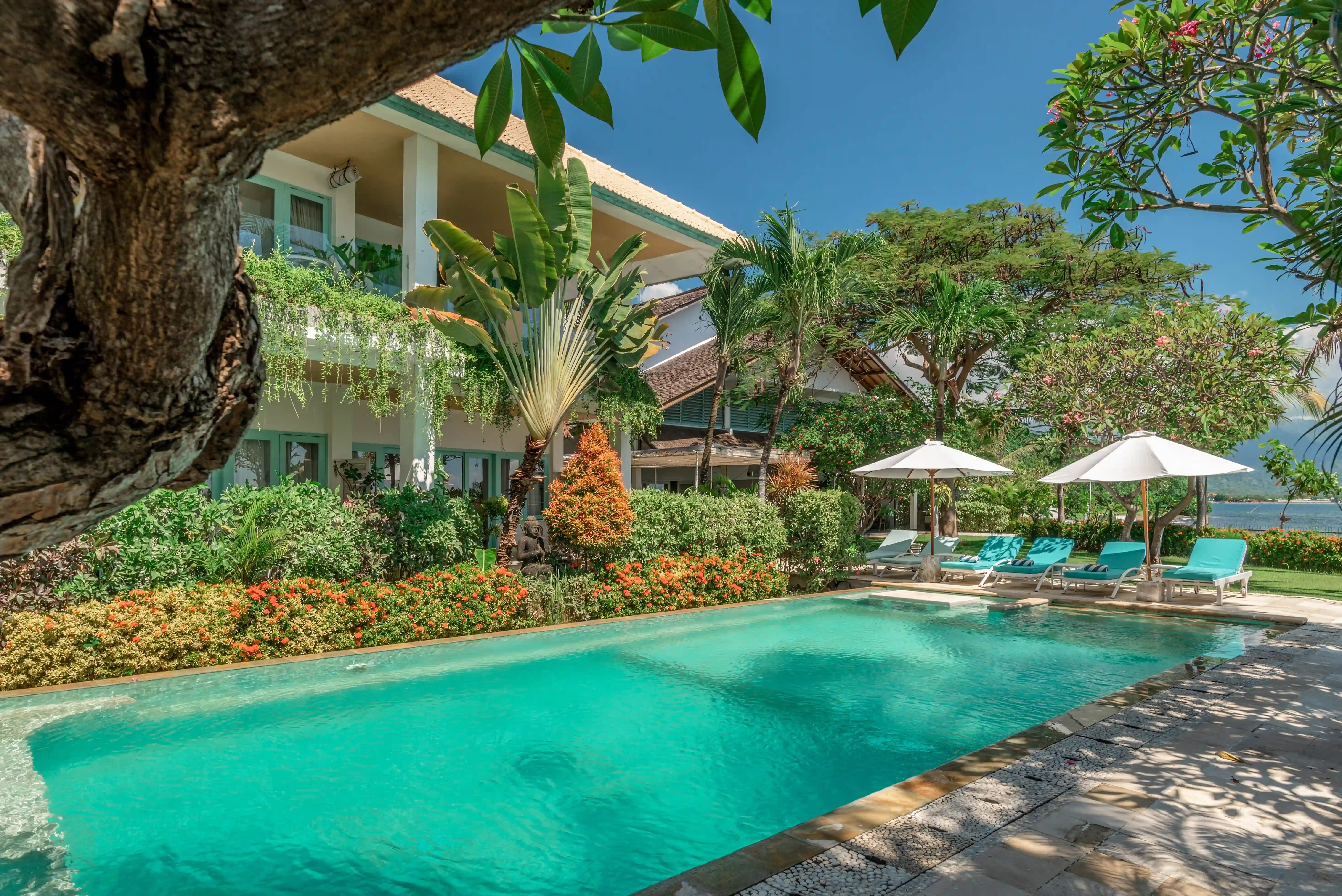 Outdoor swimming pool with sunbeds and umbrellas, with a two-story villa building in the tropical background.