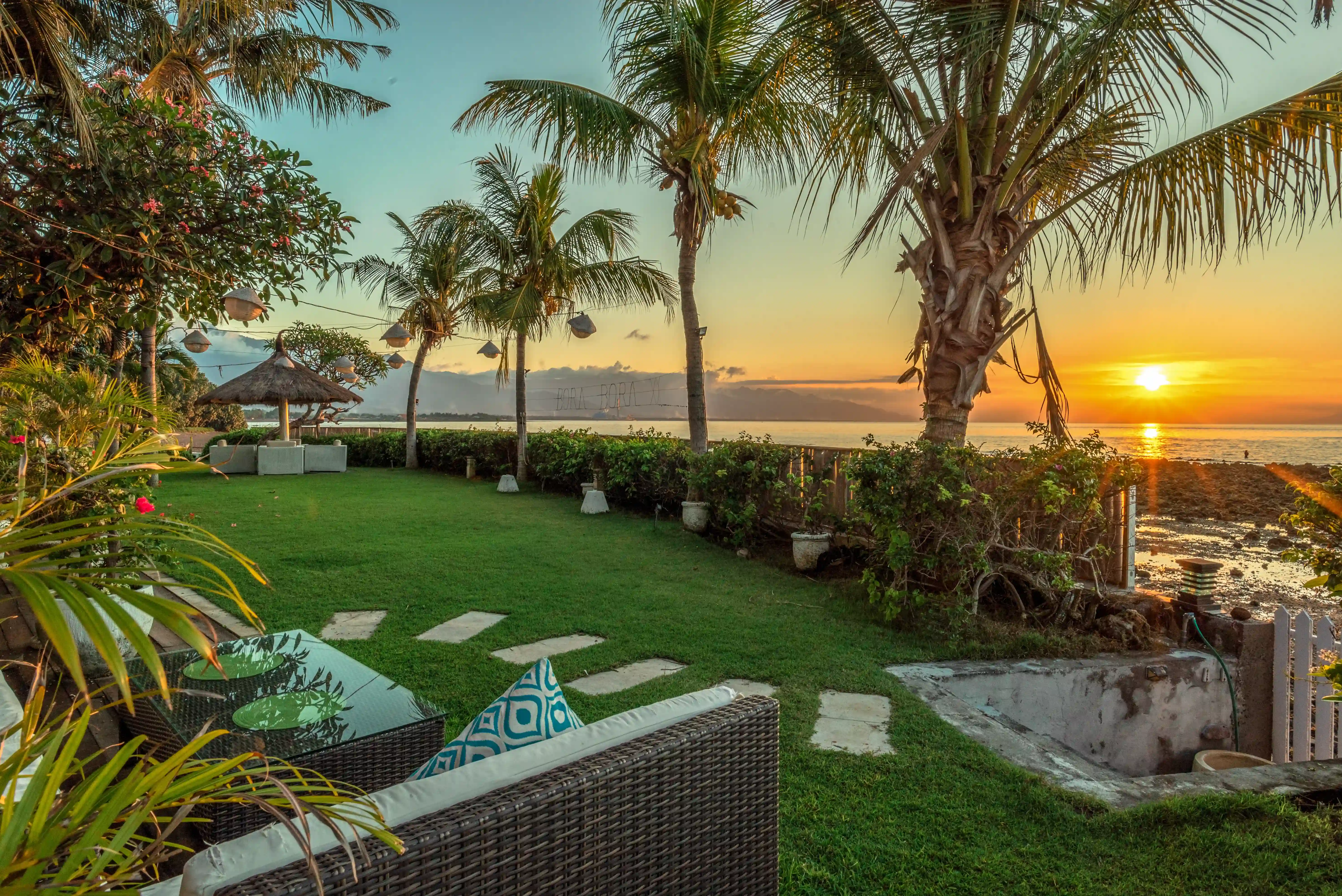 Lounge seating on the lawn with palm trees and a beautiful sunset view over the ocean, inviting guests to enjoy an evening offer.